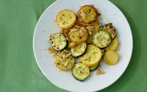 Gegrillte gelbe Zucchini-Scheiben mit Pinienkernen und Kräutern auf einem weißen Teller.