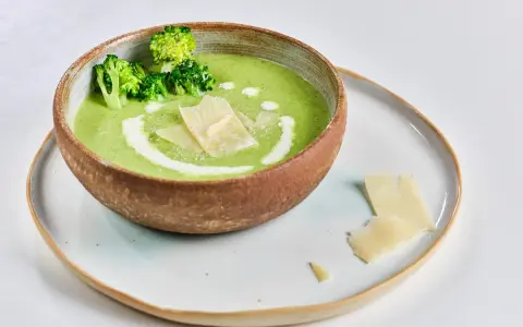 Crema de brócoli servida en un cuenco rústico, decorada con floretes de brócoli, un remolino de nata y láminas de queso parmesano.