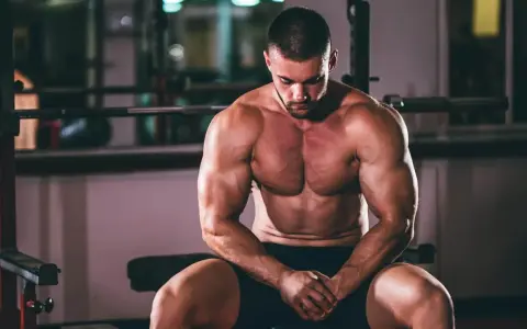 Un homme musclé, concentré, s'entraîne pour prendre du muscle rapidement. Il est assis dans une salle de sport, le regard baissé.