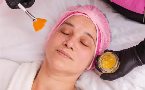 Kobieta w czepku leży z zamkniętymi oczami, a kosmetyczka przygotowuje maseczkę z miodu na twarz, używając pędzelka.