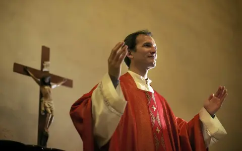 Kapłan w czerwonej szacie z podniesionymi rękami, obok krzyża z Jezusem. Wiernie przestrzega zasad prawa kanonicznego.