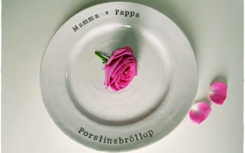 En vit tallrik med texten "Mamma ♡ Pappa" och "Porslinsbröllop". En rosa ros ligger i mitten. Ett vackert minne för de som varit gifta i 20 år.