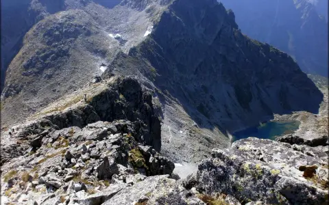 Widok z grani na skaliste zbocza i turkusowe jezioro. Mała, wysoka postać turysty na szlaku.