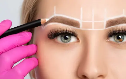 Main d'une esthéticienne en gant rose dessinant la forme sourcil visage rond avec un crayon blanc.