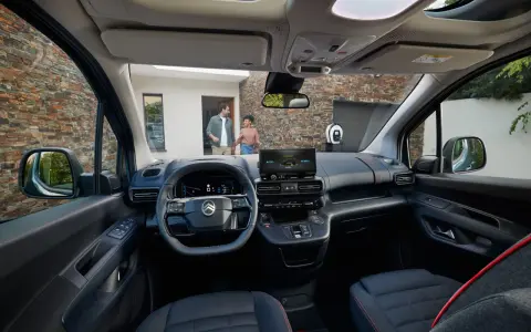 Interior de la nueva Citroën Berlingo, con pantalla táctil, volante moderno y asientos cómodos. Afuera, una pareja se acerca a un punto de carga.
