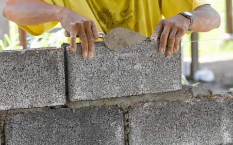 Budowa fundamentu z bloczków betonowych. Ręce murarza układają kolejny bloczek, używając kielni z zaprawą.