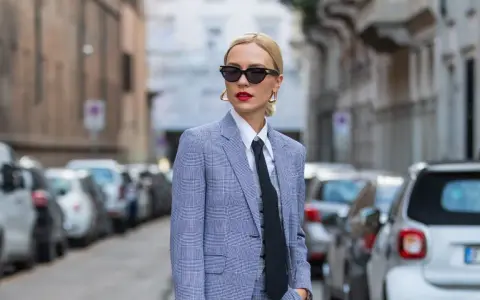 Estilo tomboy que es: mujer con traje a cuadros, camisa blanca y corbata negra, gafas de sol y labios rojos, posando en la calle.