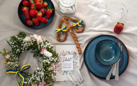 En midsommardukning med jordgubbar, blomsterkrans, en pretzel med svenska flaggor och en "Skål!"-bok. Ett personligt ljus för firandet.