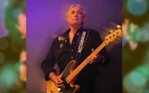 Nick Simper spielt Bassgitarre auf einer Bühne, beleuchtet von lila und grünen Lichtern.
