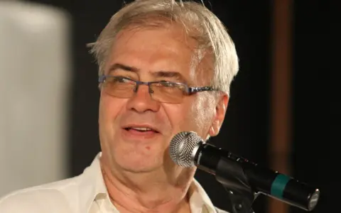 Andrzej Błaszczak, aktor, w okularach i białej koszuli, przemawia do mikrofonu.