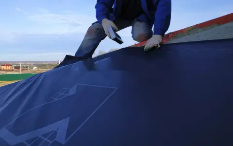 Un ouvrier en bleu, ganté, utilise un outil pour la pose d'un pare pluie sur un toit. Le ciel est bleu avec quelques nuages.