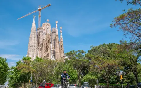 Sagrada Familia w Barcelonie, wciąż w budowie, góruje nad zielonym parkiem. Na ulicy widać rowerzystę i samochody.