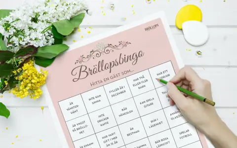 Bröllopsbingo med roliga frågor med svar. En hand med en grön penna markerar rutor på ett spelblad. Blommor ligger bredvid.