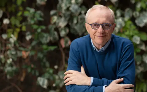 David Grossman, con occhiali blu e maglione scuro, sorride con le braccia incrociate su sfondo verde.