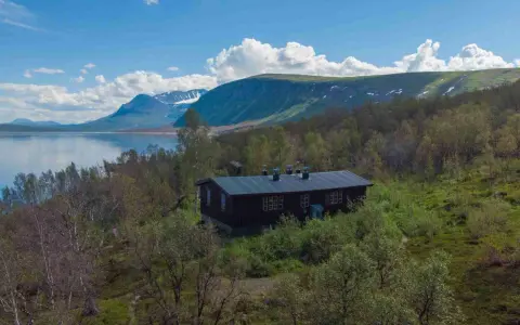 Mörkbrun byggnad vid en sjö, omgiven av björkskog och med fjäll i bakgrunden. En idyllisk plats för ritsem camping.