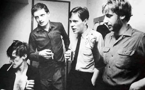 Les membres de Joy Division, Ian Curtis, Bernard Sumner, Peter Hook et Stephen Morris, posent ensemble, certains fumant, d'autres tenant des boissons.