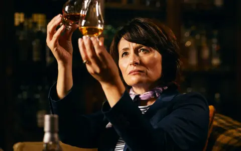 Rachel Barrie betrachtet konzentriert zwei Gläser Whisky, während sie in einer dunklen Bar sitzt.