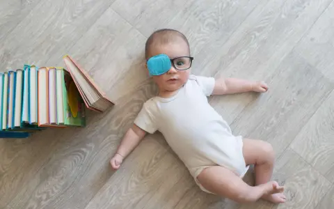 Bambino con occhiali e cerotto sull'occhio per correggere l'occhio ambliope, sdraiato vicino a una pila di libri colorati.