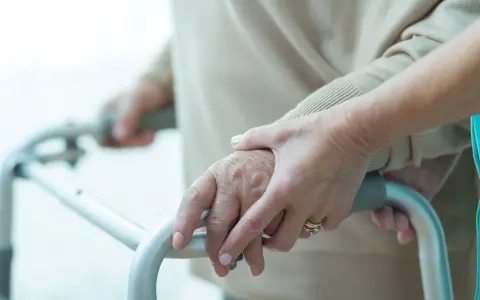 Dłonie starszej osoby i opiekuna na ramie chodzika, wsparcie przy problemach z poruszaniem się, które mogą być związane z zaburzeniami chodu ICD10.