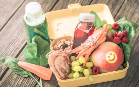 Zdrowa dieta 3 latka: żółty lunchbox z jabłkiem, malinami, winogronami, marchewką, placuszkiem i sokami.
