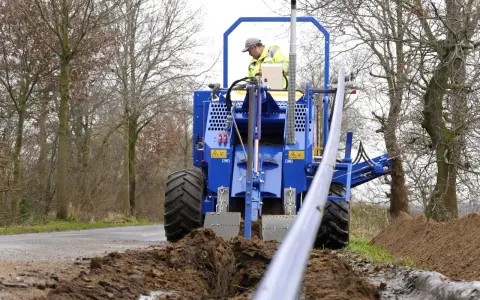 Máquina azul excava una zanja de drenaje, depositando tierra a un lado. Un operario supervisa el trabajo.