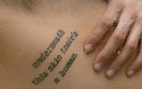 Tatuaż z cytatem "Underneath this skin there's a human" na skórze, z kroplami wody.