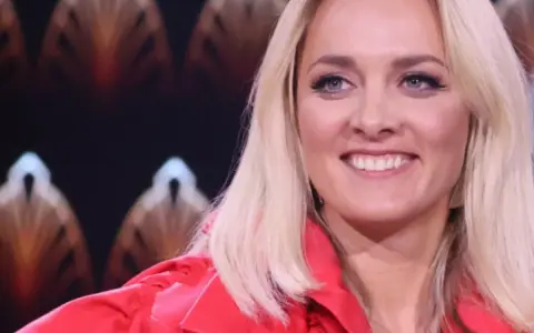 Uśmiechnięta blondynka w czerwonej marynarce, z błękitnymi oczami i delikatnym naszyjnikiem. Ola, jaka to melodia?