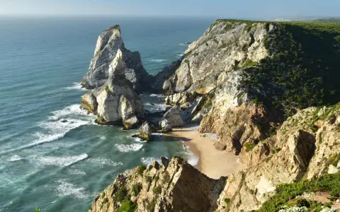 Steile Felsklippen umgeben einen einsamen sintra strand. Das türkisfarbene Meer bricht sich an den Felsen.