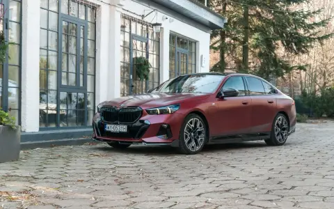 Eleganckie, matowoczerwone BMW 550e z czarnymi akcentami stoi na brukowanym podjeździe przed budynkiem z dużymi oknami.