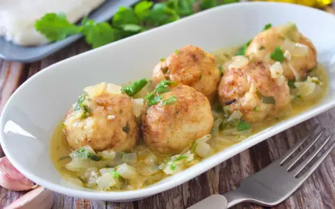 Cuatro albóndigas de merluza doradas en una salsa cremosa con cebolla y perejil, servidas en un plato blanco.