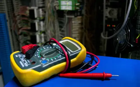 Żółty miernik cyfrowy z czerwonymi i czarnymi przewodami, gotowy do użycia. W tle widać skomplikowaną instalację elektryczną. Dowiedz się, jak używać miernika.