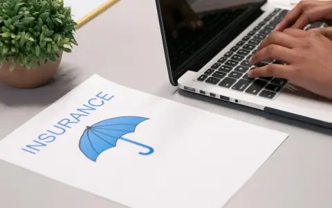 Osoba pisze na laptopie, obok dokument z napisem "INSURANCE" i parasolką. Szuka najlepszych znizki na ubezpieczenie auta.