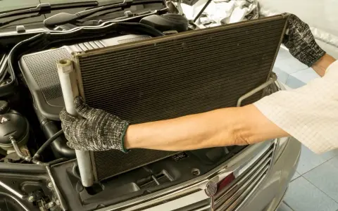 Mechanik w rękawicach wyjmuje duży, czarny skraplacz klimatyzacji z komory silnika samochodu.