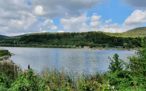 Idyllische Landschaft am Emmer-Radweg mit See, bewaldeten Hügeln und einigen Anglern am Ufer.
