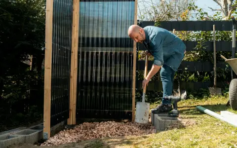 Uomo costruisce una doccia da giardino fai da te, livellando ghiaia con una pala.
