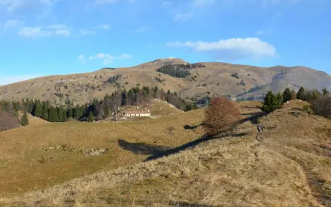 Sentieri facili sul Monte Grappa, con un escursionista che percorre un crinale erboso verso una casa isolata e la cima della montagna in lontananza.