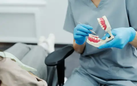 Dentysta w niebieskich rękawiczkach pokazuje na modelu, jak dbać o zęby, by uniknąć bólu dziąseł z tyłu.
