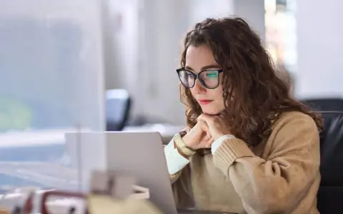 Donna con occhiali lavora al laptop, pensierosa. Potrebbe essere una dei nomadi digitali chi sono, che lavora da remoto.