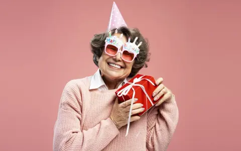 Uśmiechnięta starsza pani w okularach z napisem "Happy Birthday" i w czapce urodzinowej trzyma prezent. To dowcipne podziękowania za życzenia!