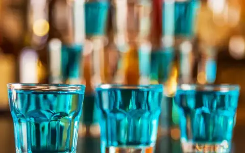 Trzy szklanki z niebieskim płynem, gotowe do wypicia jako kamikaze shot. W tle bar z butelkami.