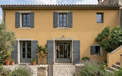 Façade d'une maison provençale à l'enduit à la chaux ocre, avec volets gris et végétation luxuriante.