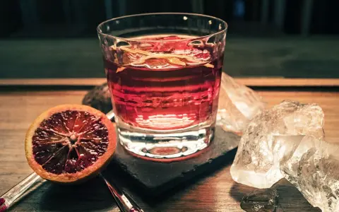 Ein Glas mit einem roten Drink, garniert mit Orangenschale, neben einer Blutorange und Eiswürfeln. Das ist Campari, ein italienischer Aperitif.