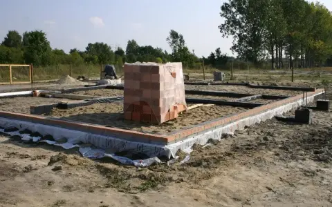 Stan zero budynku: fundamenty z cegły i betonu, piasek, folia izolacyjna, stos cegieł pod przykryciem.