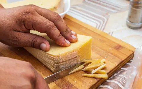 Mano cortando un bloque de queso amarillo en finas láminas. La corteza del queso se ve firme, indicando que se puede comer.