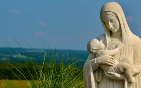 Kamienna Matka Boża ostrzega, tuląc Dzieciątko Jezus. W tle zielone wzgórza i błękitne niebo.