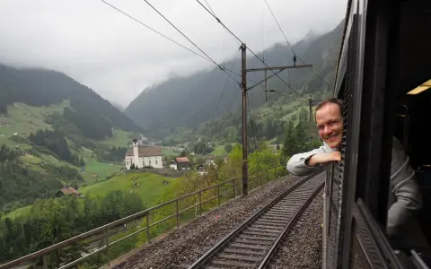 A smiling man leans out the window of the Gotthard Panorama Express, enjoying the stunning Swiss Alps scenery with a church nestled in the valley below.