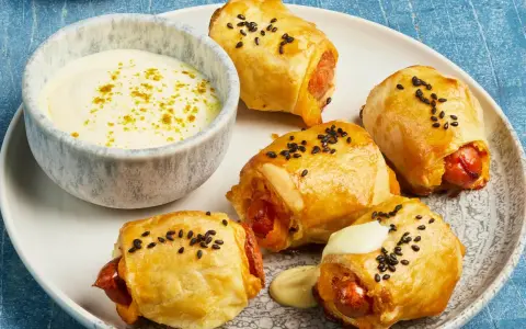 Goldbraune Blätterteigkissen mit Würstchen, bestreut mit schwarzen Sesamkörnern, serviert mit einer cremigen Dip-Sauce.