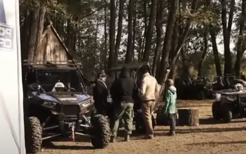 Grupa ludzi przy pojazdach terenowych podczas Polaris camping. W tle las i drewniana chata.