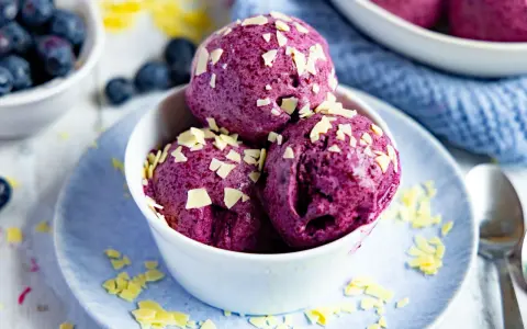 Drei Kugeln Blaubeer Eis, garniert mit weißen Schokostückchen, in einer weißen Schale auf einem blauen Teller.