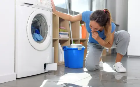 Kobieta klęczy przy pralce, zmartwiona widząc wodę pod nią. Obok stoi niebieskie wiadro.